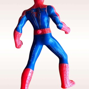 May include: A red and blue plastic action figure of Spiderman. The figure is posed with its arms outstretched and its legs spread apart. The figure has a red and blue suit with a spider logo on the chest. The figure is standing on a white background.