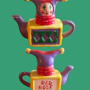 Peut inclure: Une théière en céramique en forme de bouffon avec un corps de boîte jaune, violet et vert. La théière a un chapeau de bouffon rouge et violet avec un visage peint. La théière porte les mots "RED ROSE" sur le devant.