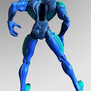 May include: Blue and green action figure with articulated limbs. The figure is posed with its back to the camera, arms bent at the elbows, and legs bent at the knees.