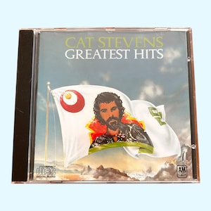 May include: A compact disc case featuring a colourful image of Cat Stevens on the front. The case is black and has the text "Cat Stevens Greatest Hits" on the front. The CD is a compilation of Cat Stevens' greatest hits.