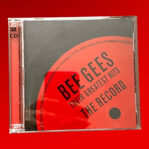 Puede incluir: Una caja de CD roja con un círculo negro en el centro. El texto "BEE GEES THEIR GREATEST HITS THE RECORD" está impreso en negro sobre el círculo rojo. El texto "2 CD" está impreso en negro en el lado izquierdo de la caja.