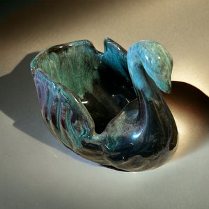 以下が含まれることがあります： 光沢のあるセラミック製の白鳥型ボウル。ボウルは、ティール、黒、バーガンディの色使いです。白鳥の頭は優雅に曲がり、ボウルの内側は広々としています。この作品はおそらく装飾用です。