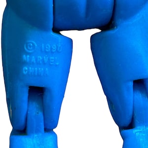 Puede incluir: Primer plano de las piernas inferiores de una figura de acci&oacute;n de pl&aacute;stico azul. Las piernas de la figura est&aacute;n dobladas por las rodillas. Los pies de la figura no son visibles. La figura tiene un s&iacute;mbolo de copyright y el texto "1990 MARVEL CHINA" impreso en la pierna izquierda.