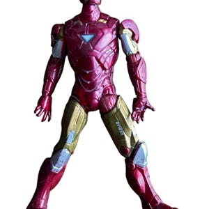 May include: Red and gold Iron Man action figure with a metallic finish. The figure is standing with its legs spread apart and its arms slightly raised. The figure has a blue and silver face mask and the text "0116IR" is printed on the right leg.
