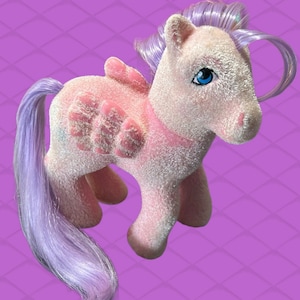 Könnte beinhalten: Ein rosa-weißes My Little Pony mit lila Mähne und Schweif. Das Pony hat ein blaues Auge und einen rosa Flügel auf dem Rücken.