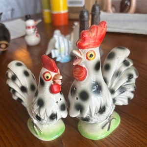 May include: A pair of ceramic rooster and hen salt and pepper shakers. The rooster and hen are white with black spots, red combs, and yellow eyes. They stand on green oval bases.