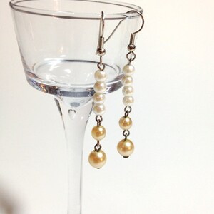 May include: A pair of dangle earrings with white and cream colored pearls. The earrings are hanging from a glass wine glass.