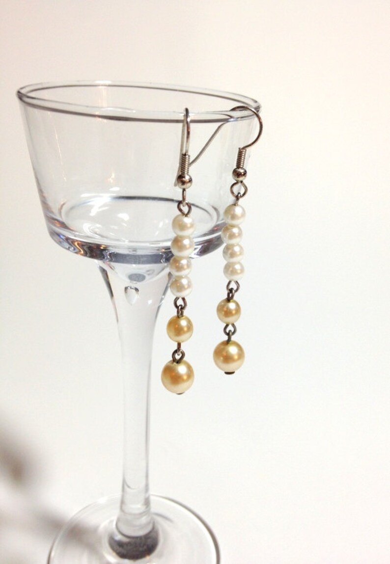 May include: A pair of dangle earrings with white and cream colored pearls. The earrings are hanging from a glass wine glass.