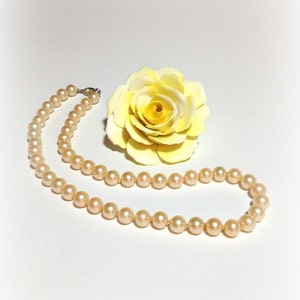 May include: A strand of cream-colored pearls on a white background with a yellow rose.