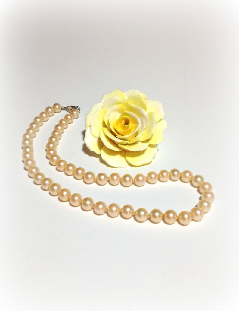 May include: A strand of cream-colored pearls on a white background with a yellow rose.