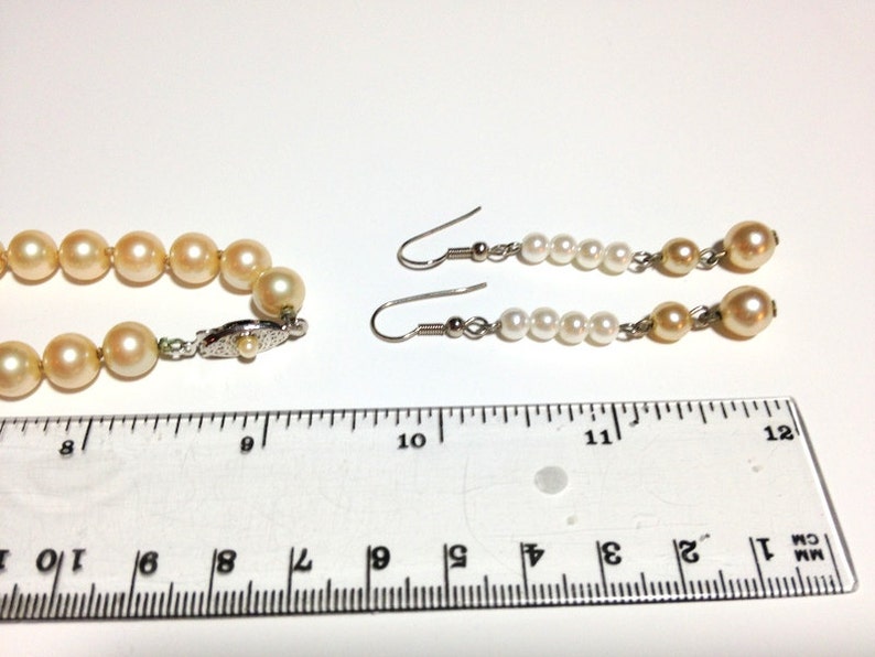May include: A pearl necklace and earrings set. The necklace is made of cream-colored pearls and has a silver clasp. The earrings are drop earrings with cream-colored pearls and silver hooks. The set is measured against a ruler.