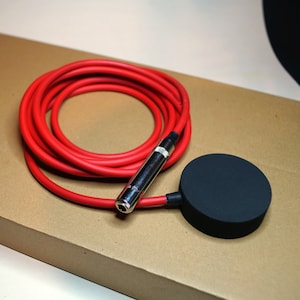 May include: A red cable coiled, attached to a silver connector and a black, circular device. The cable is thick and likely rubber. The background is a light brown surface. The device is likely a foot switch.