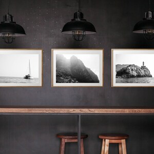 K&ouml;nnte beinhalten: Drei schwarz-wei&szlig;e gerahmte Fotografien von Meereslandschaften h&auml;ngen an einer dunkelgrauen Wand &uuml;ber einem Holztisch mit zwei Hockern. Die Fotos zeigen ein Segelboot, eine felsige Klippe und einen Leuchtturm.