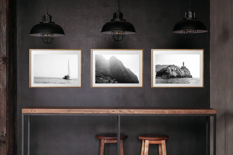 K&ouml;nnte beinhalten: Drei schwarz-wei&szlig;e gerahmte Fotografien von Meereslandschaften h&auml;ngen an einer dunkelgrauen Wand &uuml;ber einem Holztisch mit zwei Hockern. Die Fotos zeigen ein Segelboot, eine felsige Klippe und einen Leuchtturm.