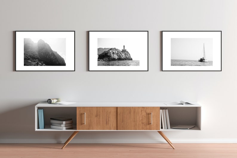 K&ouml;nnte beinhalten: Drei schwarz gerahmte Fotografien von K&uuml;stenszenen h&auml;ngen an einer wei&szlig;en Wand &uuml;ber einem wei&szlig;en Schrank aus Holz mit Regalen. Das linke Foto zeigt eine felsige Bergseite mit Sonnenschein, der durch die Wolken scheint. Das mittlere Foto zeigt eine felsige Insel mit einem Leuchtturm auf der Spitze. Das rechte Foto zeigt ein Segelboot auf dem Wasser.