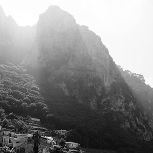K&ouml;nnte beinhalten: Ein Schwarzwei&szlig;foto eines felsigen Berghangs mit einem kleinen Dorf am Fu&szlig;e. Der Berg ist mit Nebel bedeckt und das Dorf ist kaum zu sehen.
