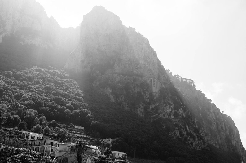 K&ouml;nnte beinhalten: Ein Schwarzwei&szlig;foto eines felsigen Berghangs mit einem kleinen Dorf am Fu&szlig;e. Der Berg ist mit Nebel bedeckt und das Dorf ist kaum zu sehen.