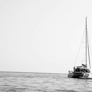 K&ouml;nnte beinhalten: Ein wei&szlig;es Segelboot mit einem einzelnen Mast und Segeln liegt in der Mitte eines ruhigen, grauen Ozeans vor Anker. Der Himmel ist klar und hell.