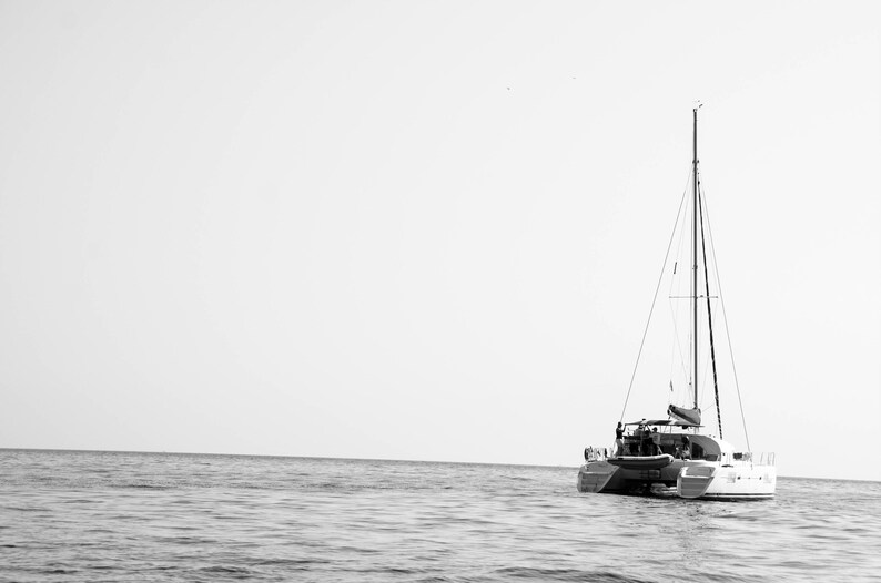 K&ouml;nnte beinhalten: Ein wei&szlig;es Segelboot mit einem einzelnen Mast und Segeln liegt in der Mitte eines ruhigen, grauen Ozeans vor Anker. Der Himmel ist klar und hell.