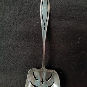 May include: A silver-toned metal serving utensil featuring a wide, square-shaped head with decorative cutouts and a long, slender handle. The handle includes an elongated, pointed tip with symmetrical openwork detailing and a small engraved monogram.