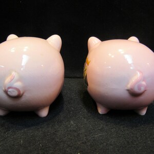 May include: Two pink ceramic piggy banks, viewed from the rear, showcasing their curly tails and small legs. One pig has a decorative design on its side. The banks are on a dark surface.