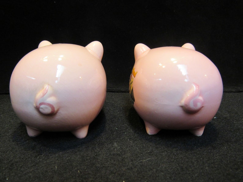 May include: Two pink ceramic piggy banks, viewed from the rear, showcasing their curly tails and small legs. One pig has a decorative design on its side. The banks are on a dark surface.