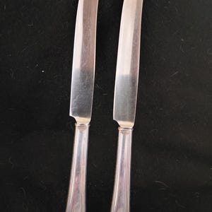 May include: Two silver butter knives with a decorative handle design. The blades are curved and the handles have a detailed, ridged pattern. The knives are set against a dark background, highlighting their polished appearance.