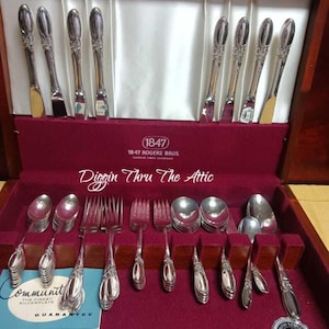 Puede incluir: Un juego de cubiertos antiguos en una caja de madera. El juego incluye cuchillos, tenedores y cucharas con mangos ornamentados. La caja tiene un forro de terciopelo burdeos. El texto "1847 Rogers Bros" es visible.