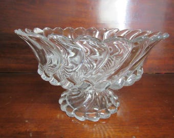 May include: A clear glass bowl with a scalloped edge and a swirl design. The bowl is on a pedestal base.