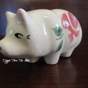 May include: White ceramic piggy bank with a pink rose and green leaves painted on the side. The piggy bank has black eyes and a small mouth.
