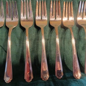 Puede incluir: Siete tenedores plateados con púas y mangos alargados están dispuestos sobre un paño verde. Los tenedores tienen un aspecto metálico brillante, que refleja la luz. Los mangos tienen un diseño geométrico decorativo.