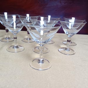 May include: Set of six clear glass martini glasses. Each glass has a wide, triangular bowl and a slender stem with a decorative element. The glasses are arranged on a light-colored surface, suggesting a formal setting.