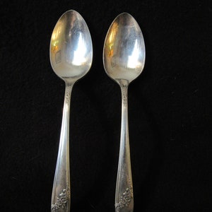 May include: Two silver spoons with ornate floral designs on the handles. The spoons are on a black background.