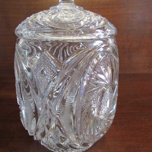 May include: A clear glass jar with a lid, featuring intricate cut glass designs. The jar has a round body and a domed lid with a knob on top. The glass is clear and reflects light.