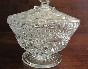 May include: A clear pressed glass candy dish with a matching lid. The bowl and lid feature a repeating diamond pattern and a scalloped rim. The lid is topped with a pointed, faceted finial, and the base is a circular pedestal with a sunburst design.