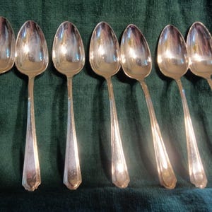 Puede incluir: Siete cucharas de plata con cuencos ovalados y mangos largos facetados están dispuestas sobre un paño verde oscuro. Las cucharas reflejan la luz, creando una apariencia brillante y brillante. Las cucharas son probablemente para servir o comer.