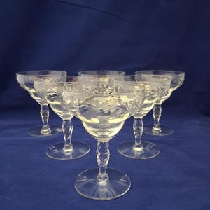 May include: Six clear, stemmed champagne glasses with etched floral designs. The glasses have a rounded bowl, a slender stem, and a circular base. The glasses are arranged on a blue background.