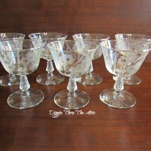 Puede incluir: Juego de ocho vasos de cristal transparente con tallo y diseño floral grabado. Los vasos están dispuestos en círculo sobre una superficie de madera.