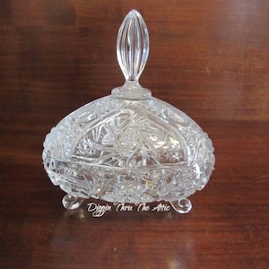 May include: A clear glass candy dish with a lid and three legs. The dish is decorated with a cut glass pattern.