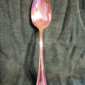 Puede incluir: Una cuchara plateada con una superficie reflectante. La cuchara tiene un cuenco ovalado y un mango largo y delgado que se ensancha en la base. El mango tiene un diseño decorativo y parece ser de metal. La cuchara está sobre un fondo verde oscuro.
