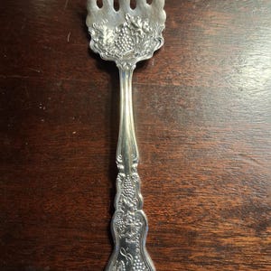 May include: A silver-toned metal serving fork featuring an ornate handle and tines with a decorative embossed pattern of grape clusters and vine leaves. The handle includes an engraved script letter S near the base, with a polished metallic finish.