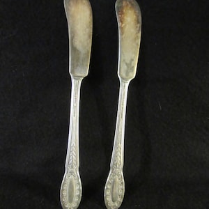 May include: Two silver butter knives with ornate handles. The knives have a slightly curved blade and a rounded tip.