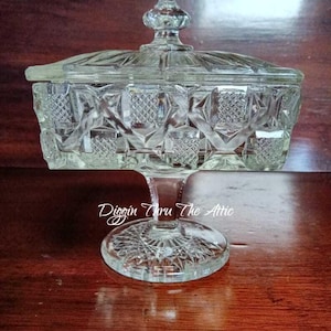 May include: A clear, vintage glass candy dish with a lid. The dish is rectangular with diamond-shaped patterns and sits on a pedestal base. The lid has a decorative finial. The text "Diggin Thru The Attic" is visible on the base.