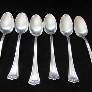 Puede incluir: Seis cucharaditas de plata con mangos ornamentados. Las cucharas tienen un acabado pulido y están dispuestas en fila.