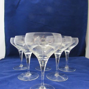 May include: A set of five clear glass coupe glasses with wide, shallow bowls and slender stems. Each glass features a smooth, transparent finish and a circular base, arranged in a cluster to showcase the uniform design of the glassware.