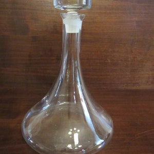 May include: A clear glass decanter with a round, wide base and a tall, slender neck. The decanter has a clear glass stopper with a flat top.