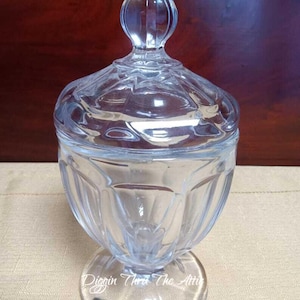 Puede incluir: Un cuenco de cristal transparente con tapa, sobre un pedestal. El cuenco tiene una base redonda, un cuerpo bulboso y facetado, y una tapa con un remate en forma de lágrima. El texto "Diggin Thru The Attic" es visible en la base.