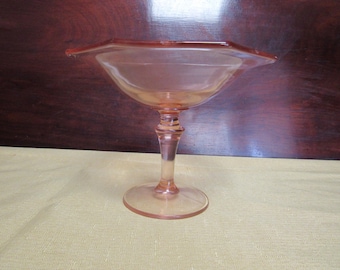 May include: A vintage, pink-colored glass pedestal bowl. The bowl has a hexagonal rim and sits atop a stem and circular base. The glass has a smooth, transparent appearance. The bowl is likely used for serving desserts or displaying decorative items.