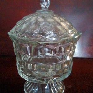 Puede incluir: Un cuenco de cristal transparente para caramelos con tapa. El cuenco tiene un patrón geométrico texturizado y se asienta sobre una base de pedestal. La tapa tiene un pequeño asa. El texto "Diggin Thru The Attic" está en la parte inferior.