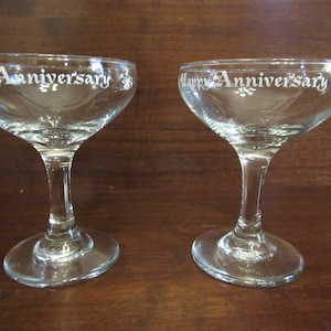 May include: Two clear glass champagne coupes with the words "Happy Anniversary" etched on the front of each glass.